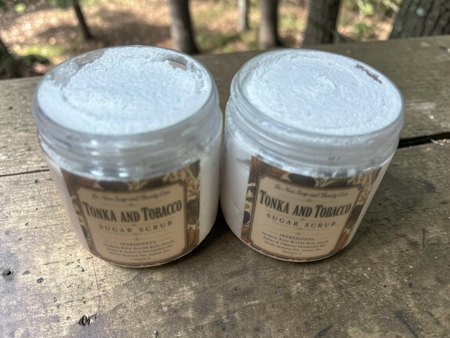 La Neve Soap and Beauty Care Tonka and Tobacco foaming sugar scrub in two clear jars on a rustic wooden surface.