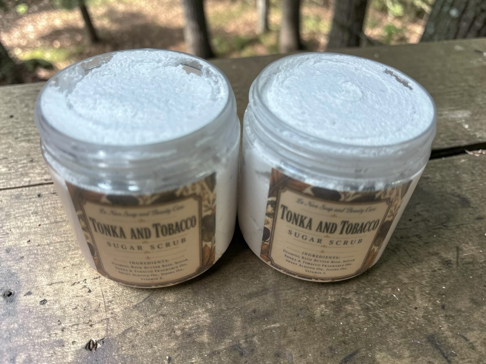 La Neve Soap and Beauty Care Tonka and Tobacco foaming sugar scrub in two clear jars on a rustic wooden surface.