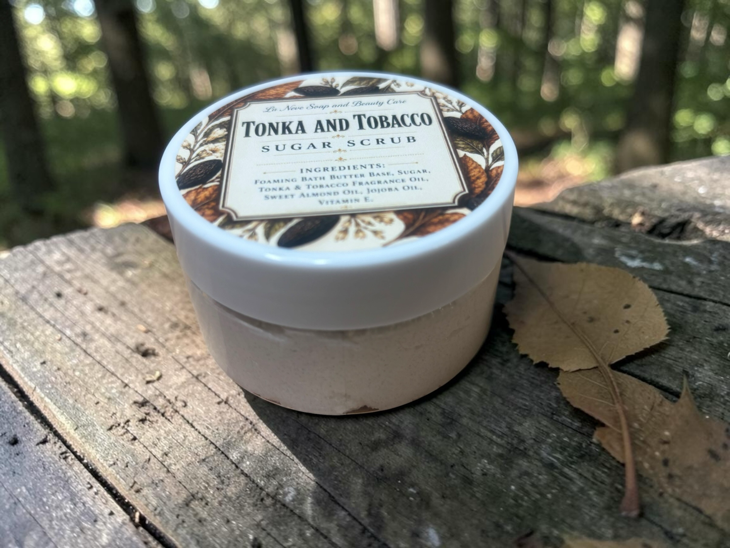La Neve Soap and Beauty Care Tonka and Tobacco sugar scrub in a white jar on a wooden surface in a forest setting.