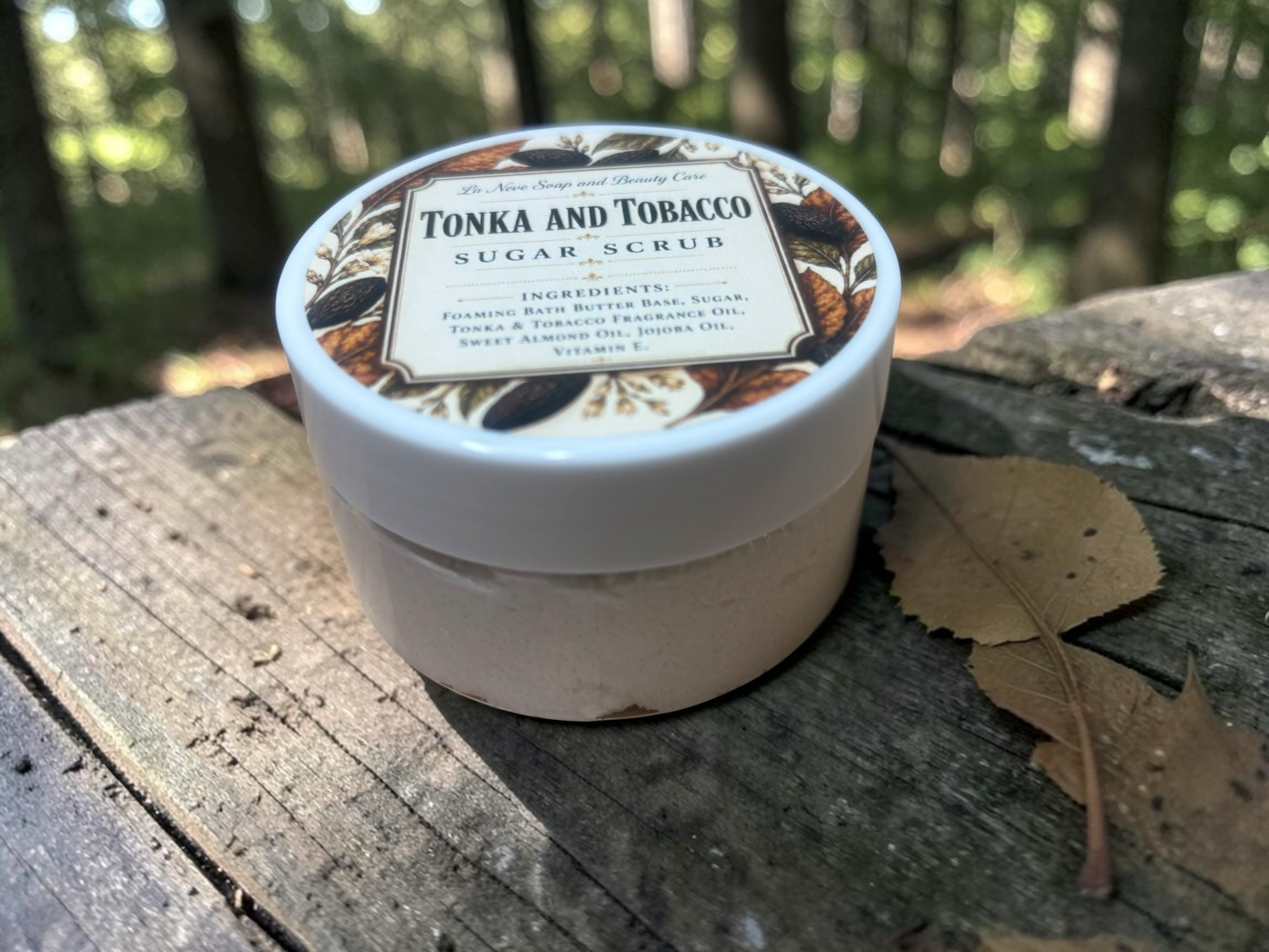 La Neve Soap and Beauty Care Tonka and Tobacco sugar scrub in a white jar on a wooden surface in a forest setting.