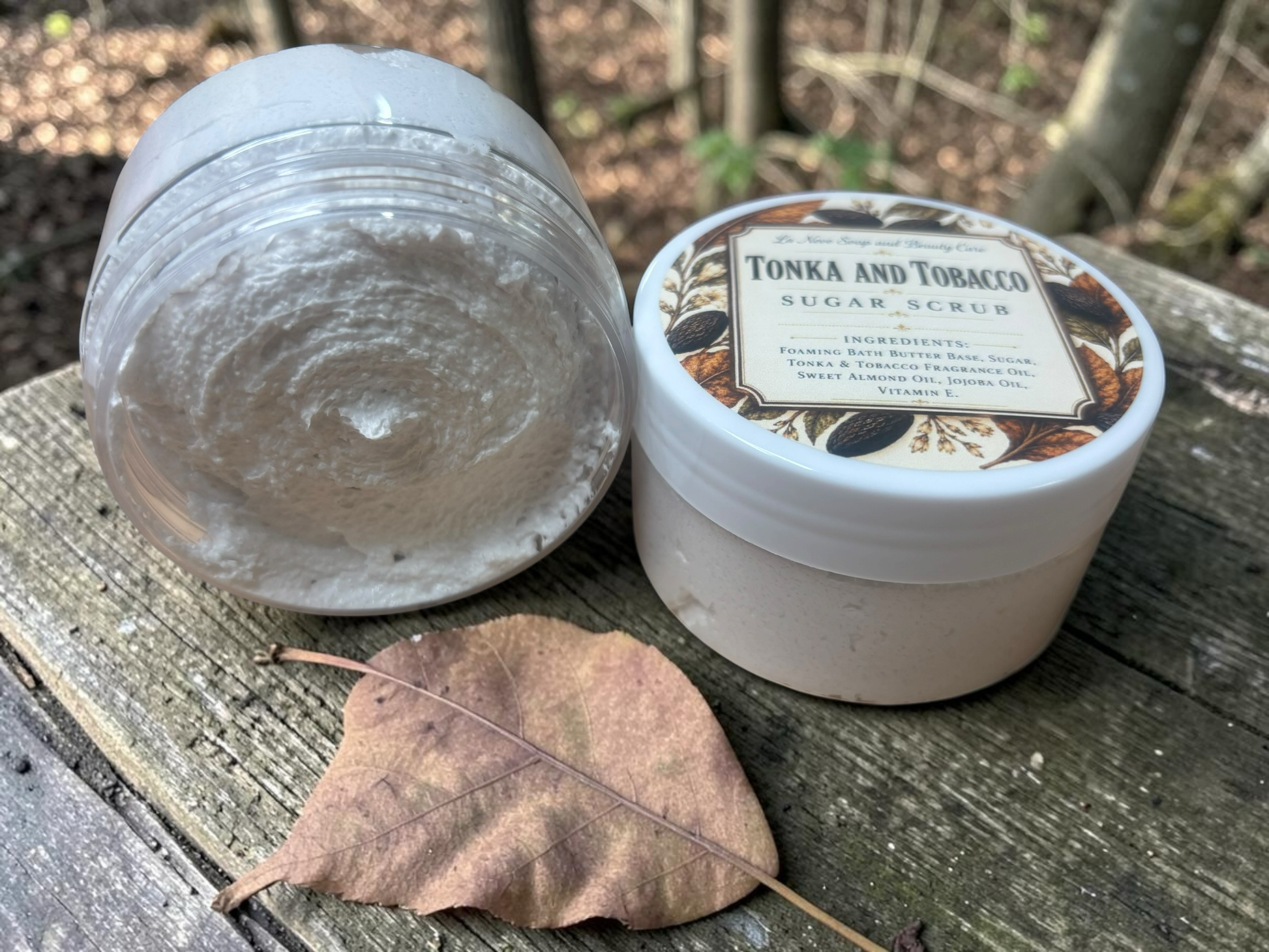 La Neve Soap and Beauty Care Tonka and Tobacco foaming sugar scrub in a jar on a wooden surface with a dry leaf.