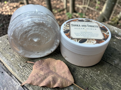 La Neve Soap and Beauty Care Tonka and Tobacco foaming sugar scrub in a jar on a wooden surface with a dry leaf.