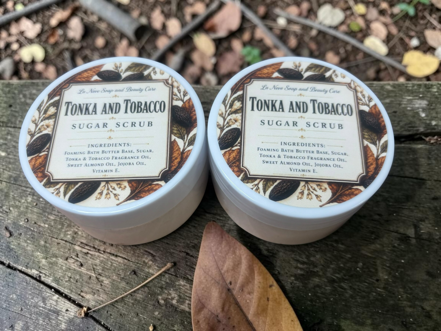 La Neve Soap and Beauty Care Tonka and Tobacco sugar scrub jars on a rustic wooden surface with autumn leaves.