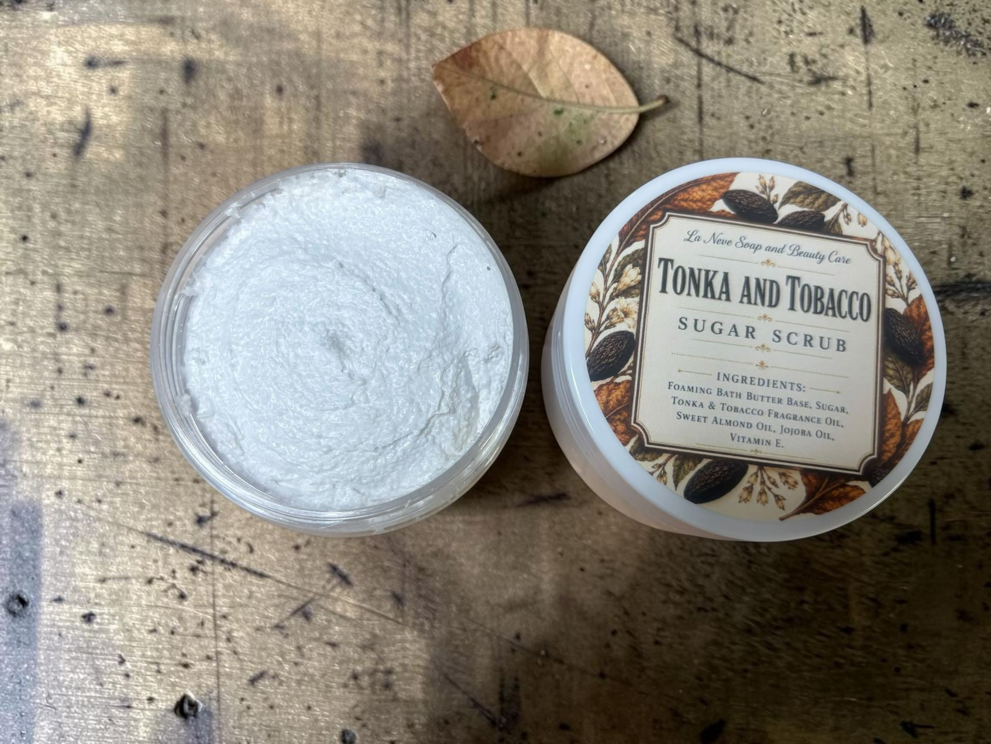 La Neve Soap and Beauty Care Tonka and Tobacco sugar scrub in a jar with a decorative label on a rustic wooden table.